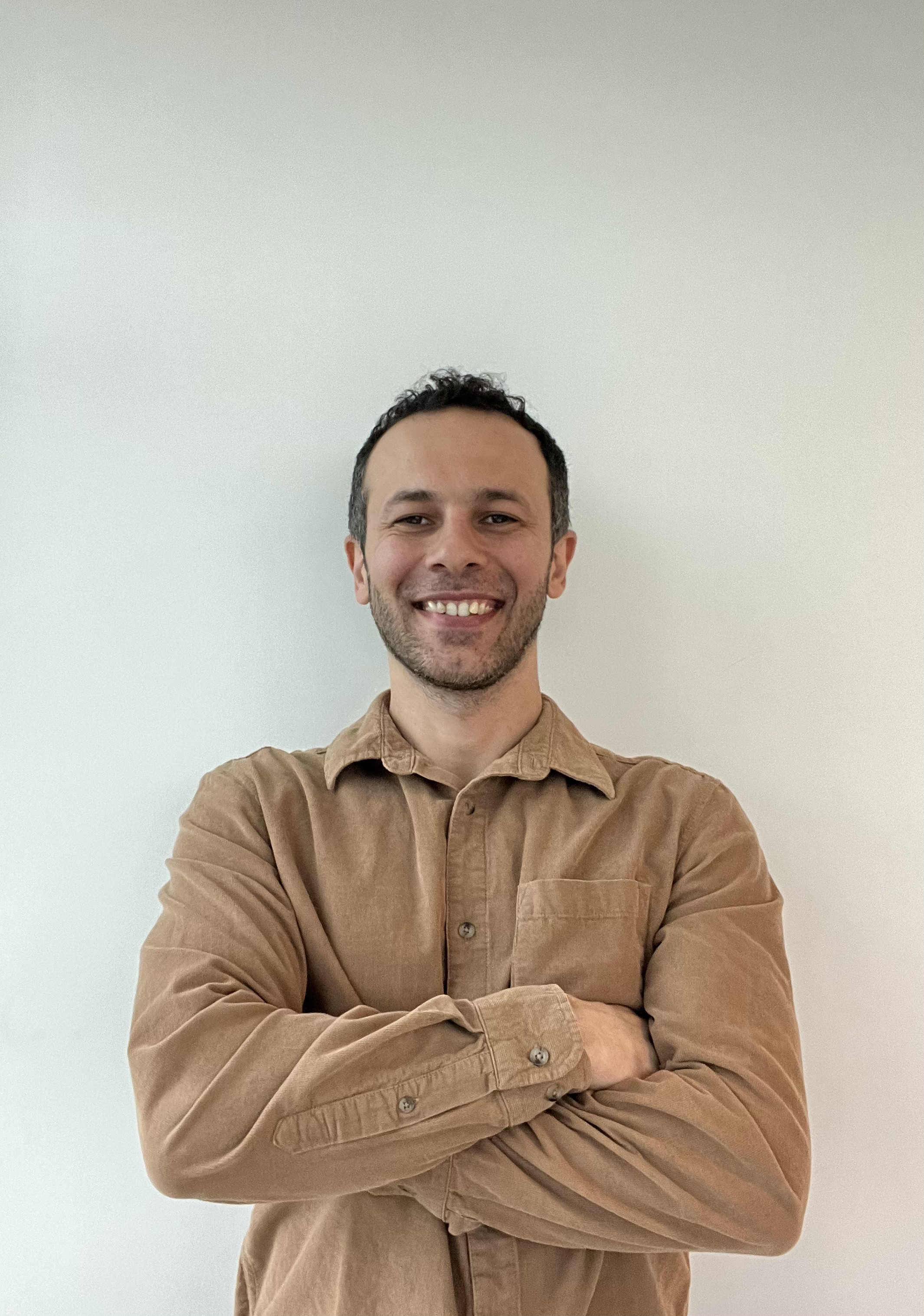 Man smiling with arms crossed, standing in front of a plain light-colored wall and wearing a beige shirt.